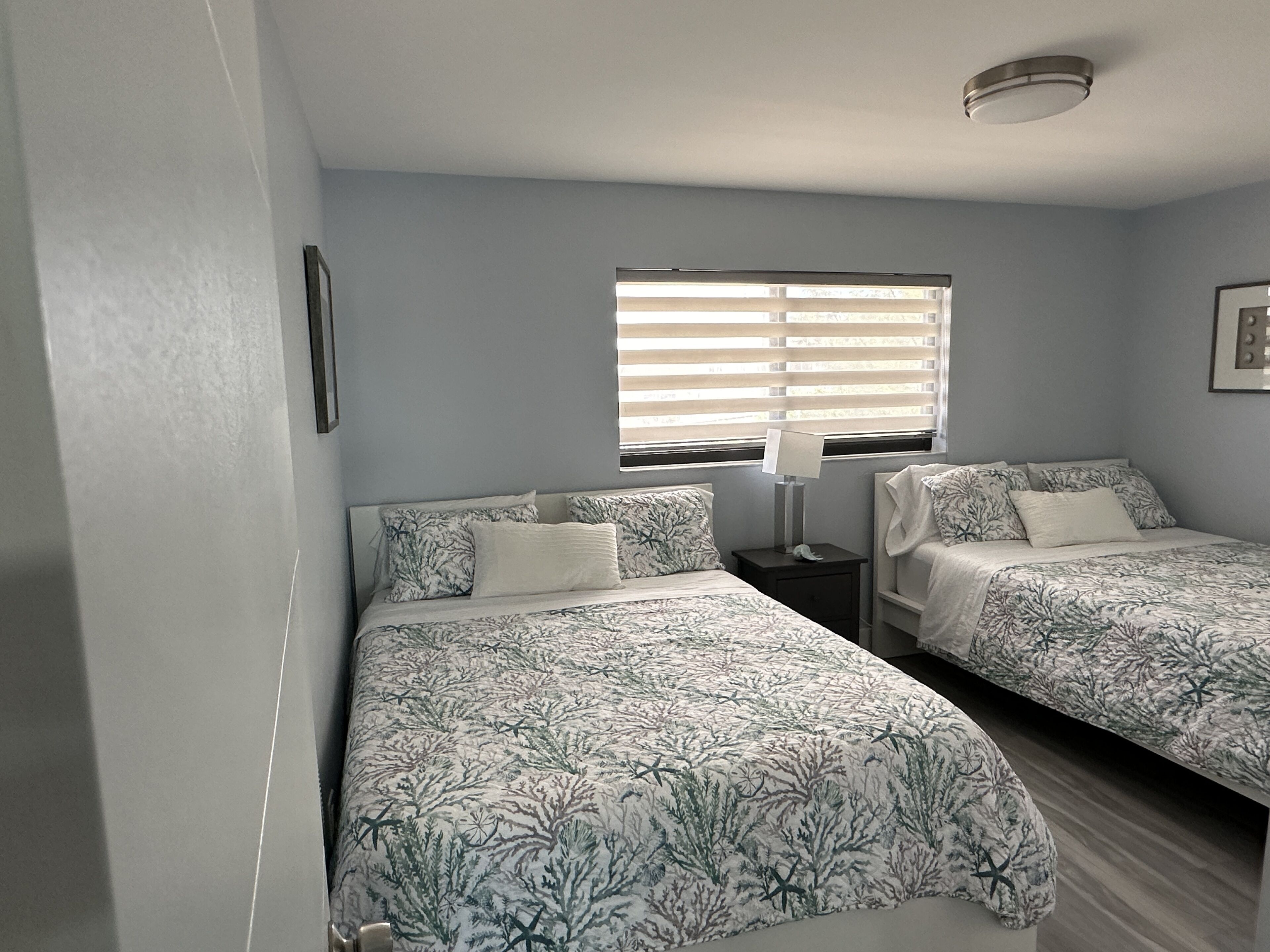 A bedroom with two beds, a window with blinds, a bedside table with a lamp, and a framed picture on the wall.