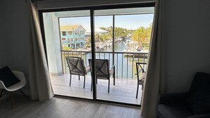 A room with a large sliding glass door leading to a balcony with two chairs and a table. The balcony overlooks a body of water with boats and buildings in the background. - Re-energize at our spacious condo! Get 10% off stays of 7+ days. Book now! (Key Largo)