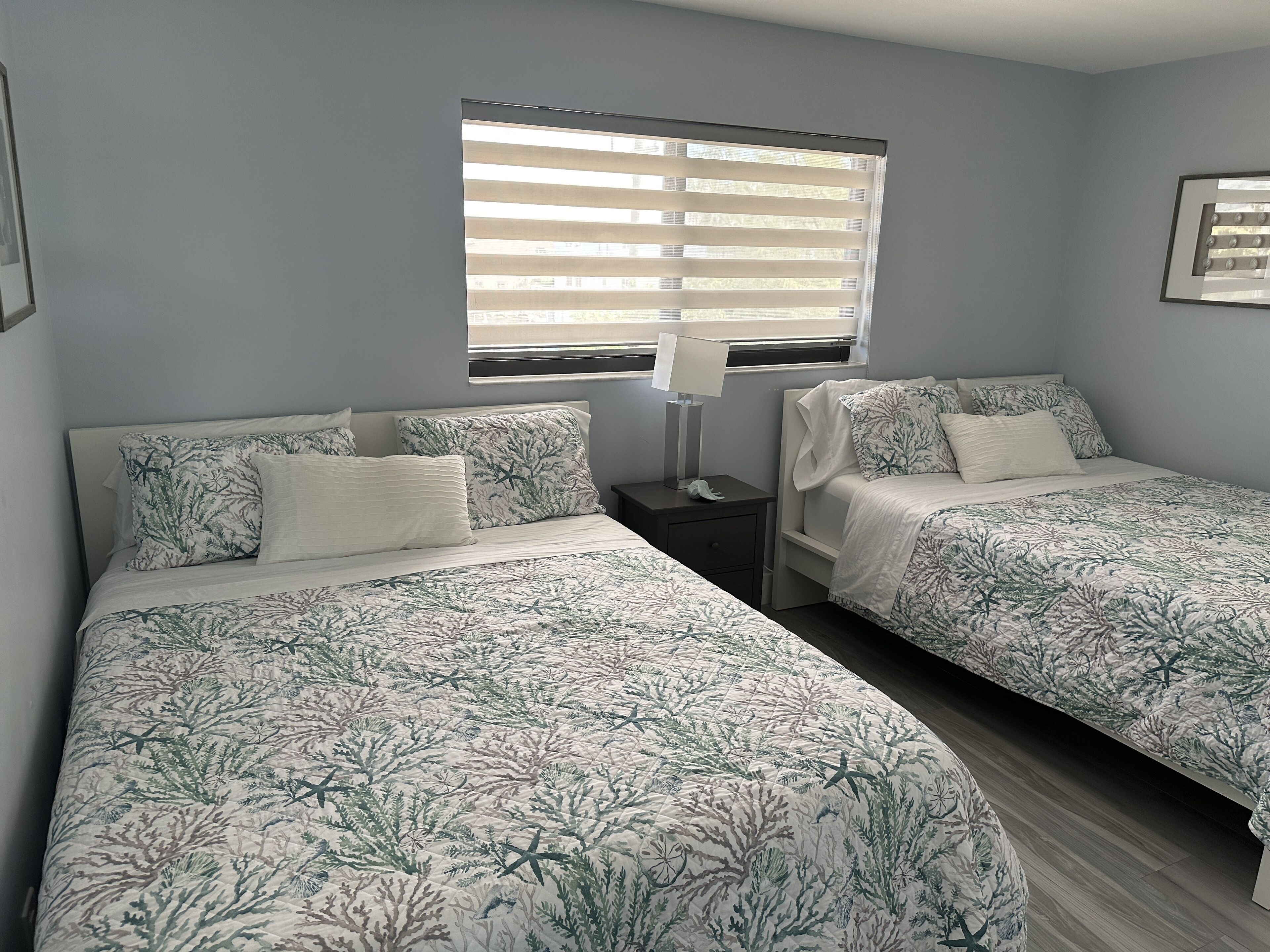 A bedroom with two beds, each with a patterned bedspread, a bedside table with a lamp, and a window with blinds.