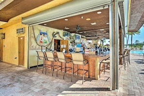A bar area with a wooden counter and multiple bar stools. There are ceiling fans and a pool visible in the background.