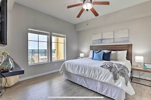 A bedroom with a bed, a ceiling fan, a window with a view, and a nightstand with a lamp.