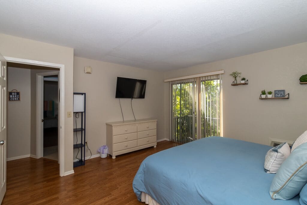 A bedroom with a bed, a dresser, a television, and a sliding glass door to a balcony.