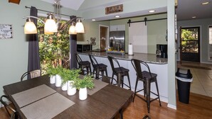 A modern kitchen with a central island, bar stools, and a dining table with placemats and potted plants.