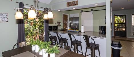 A modern kitchen with a central island, bar stools, and a dining table with placemats and potted plants.