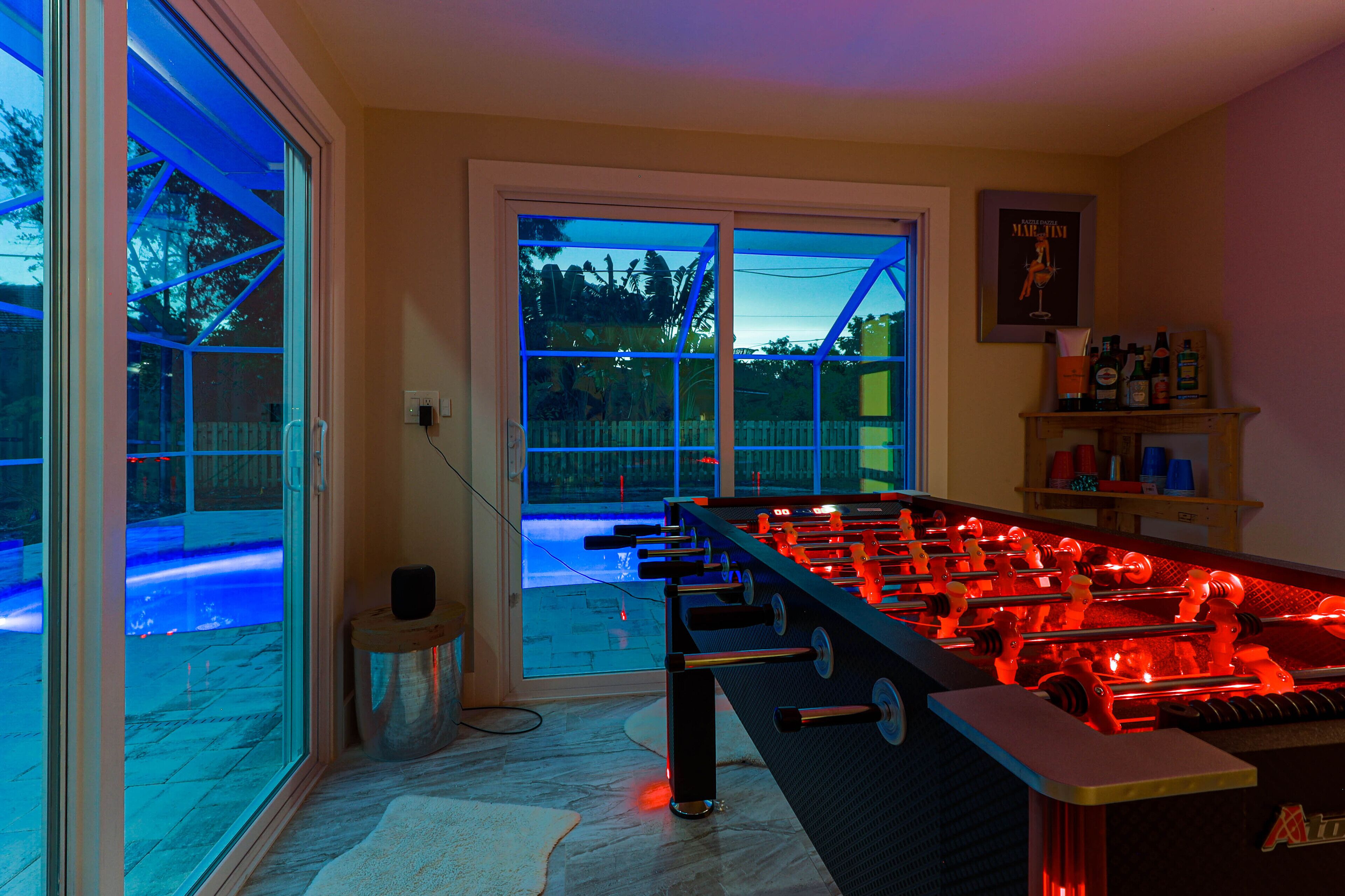 A foosball table in a room with a large window, a door leading outside, and a shelf with bottles.