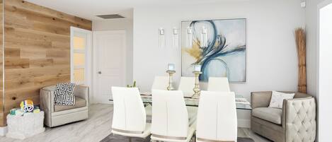 A modern living room with a dining area, featuring a wooden accent wall, a large abstract painting, and a glass dining table with white chairs.