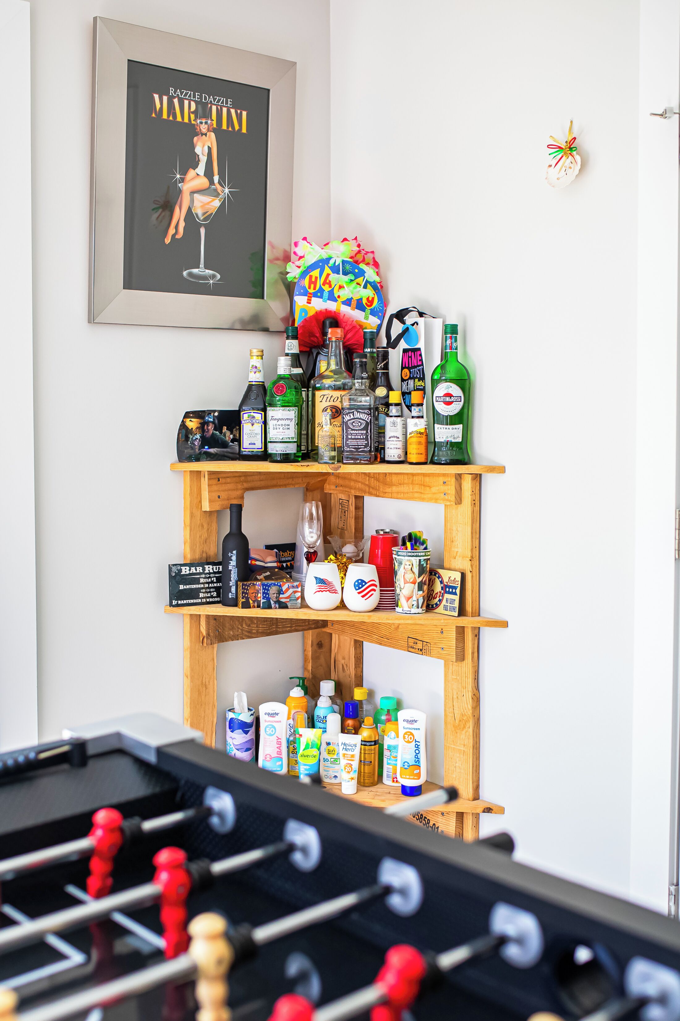 A room with a foosball table, a shelf with various bottles and a framed picture on the wall.
