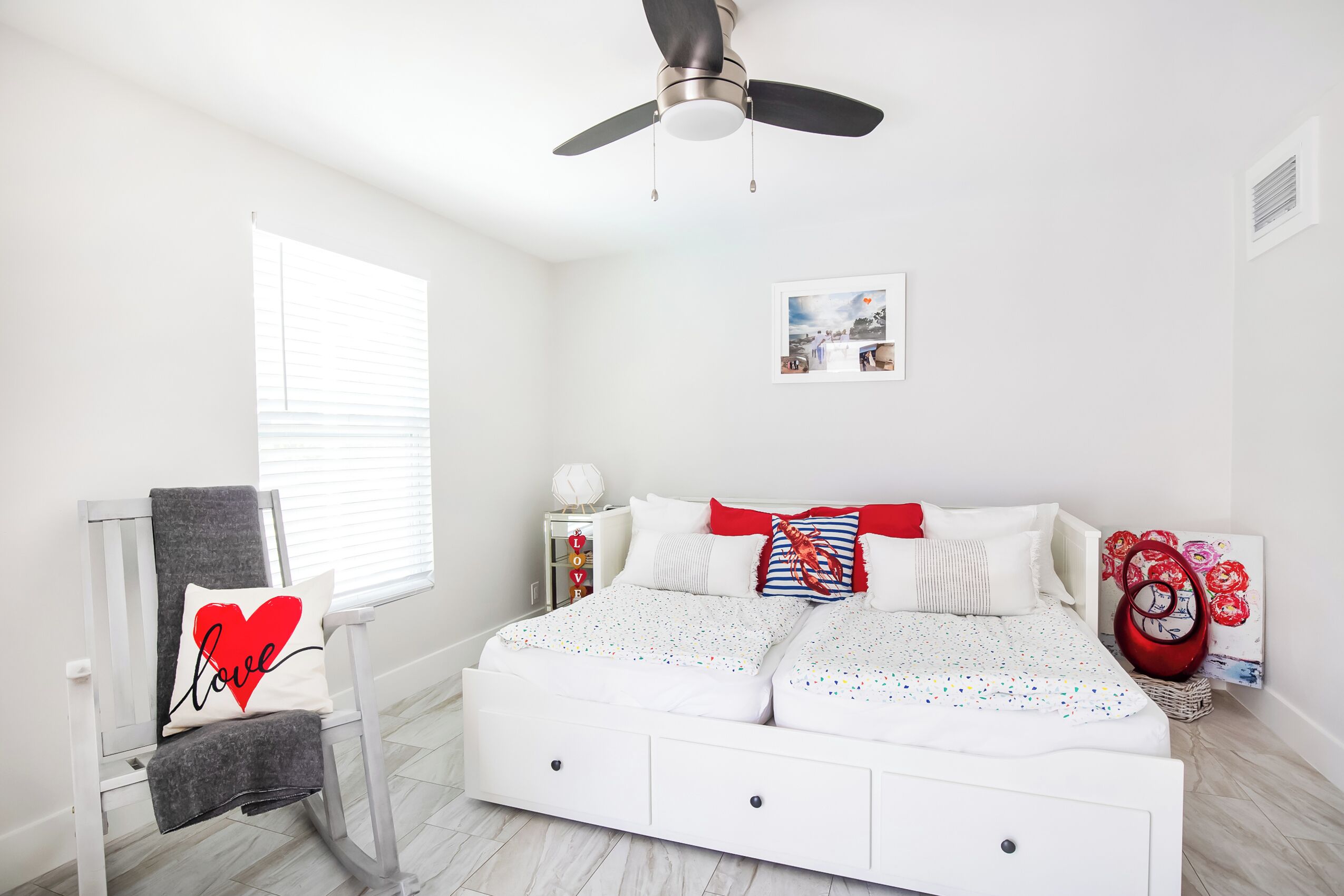 A bedroom with a white bed, a chair with a towel, a ceiling fan, a bedside table, and a window with blinds.