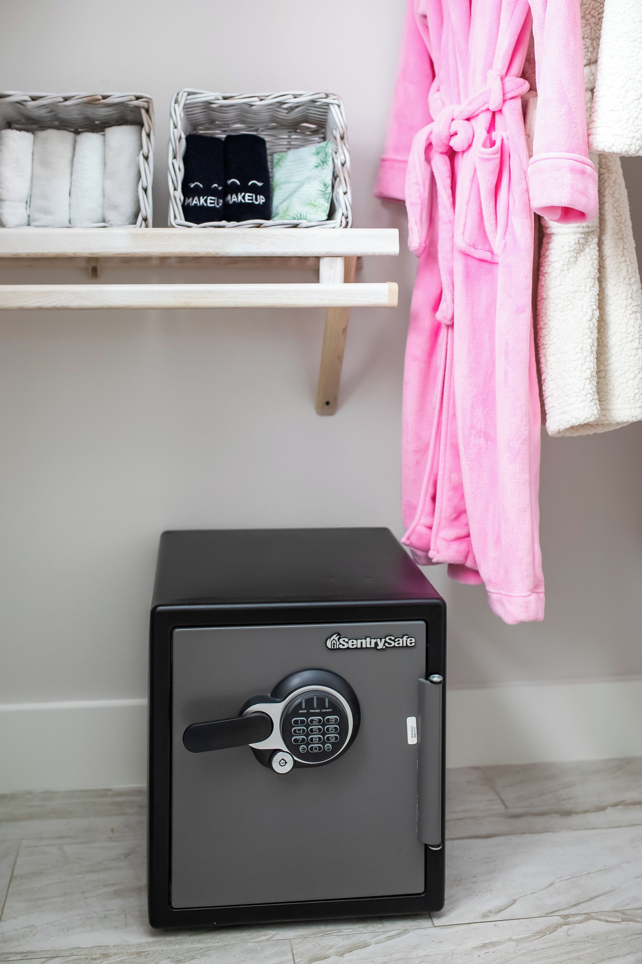 A black safe with a keypad lock, a pink robe, and a shelf with folded towels and a black item labeled "MAKEUP".
