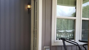 Un porche cubierto con piso de madera, una ventana con vista a una casa y una silla de metal.