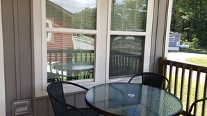 Una mesa de vidrio con cuatro sillas sobre una terraza de madera, con vistas a un patio cercado con árboles.