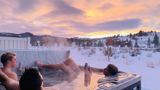 Una vasca idromassaggio con tre persone che si rilassano, immersa in un paesaggio innevato e illuminata da un tramonto.