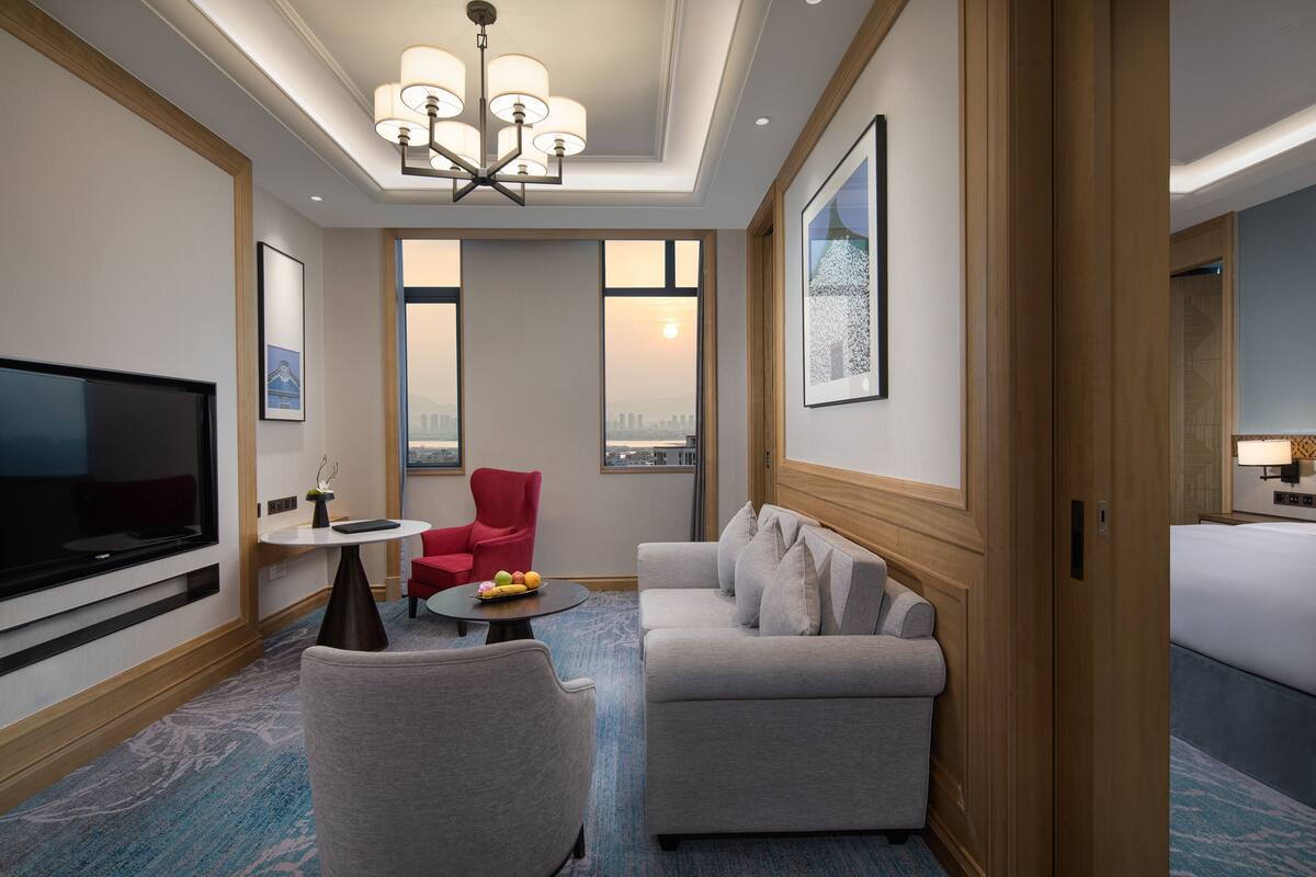 Een hotelkamer met een grote televisie, een rode fauteuil, een grijze bank, een kleine tafel met fruit en een stadszicht door de ramen.