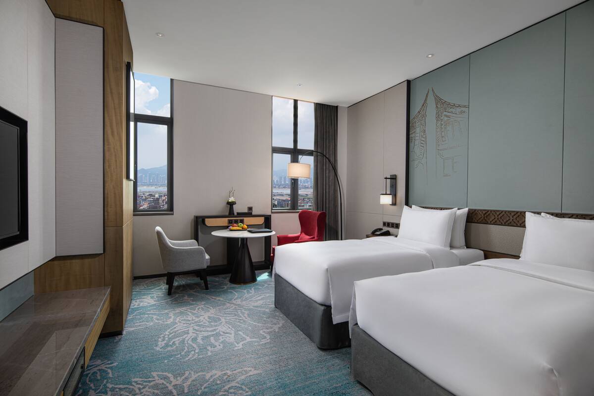 Een hotelkamer met twee bedden, een bureau, een stoel, een televisie en uitzicht op de stad.