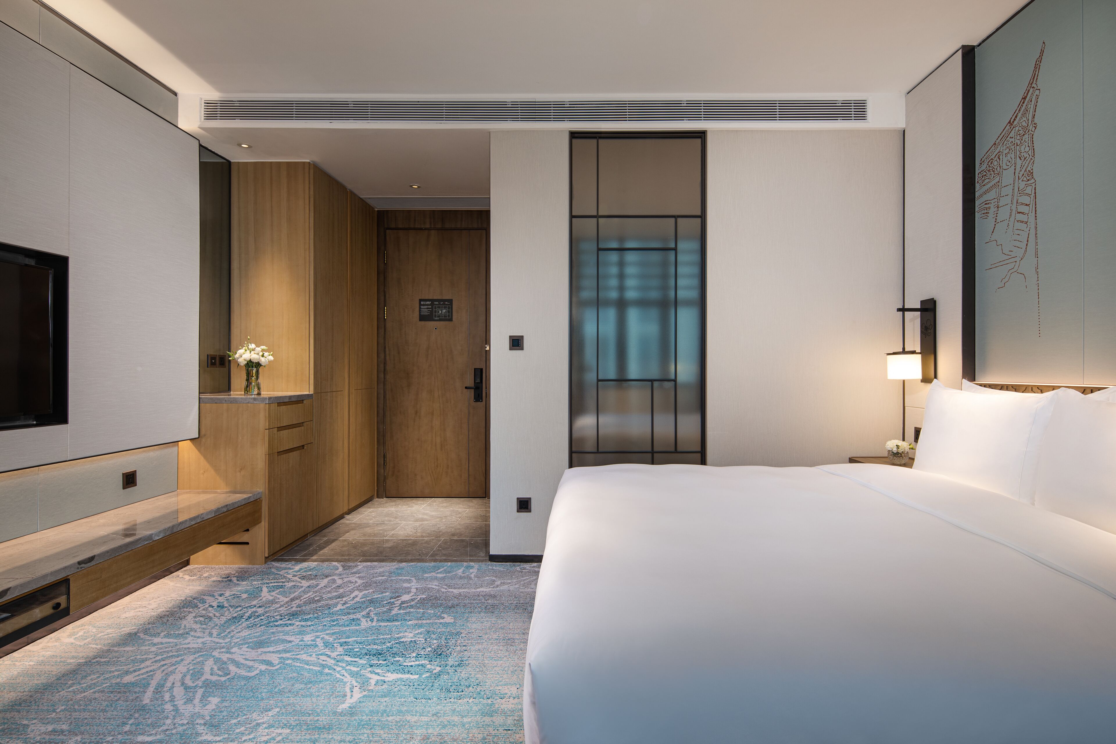 Quarto de hotel com uma cama grande, televisão, porta de madeira e vista para a cidade através de uma janela.