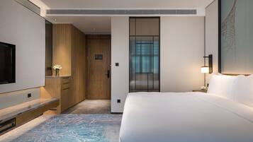 Quarto de hotel com uma cama grande, televisão, porta de madeira e vista para a cidade através de uma janela.