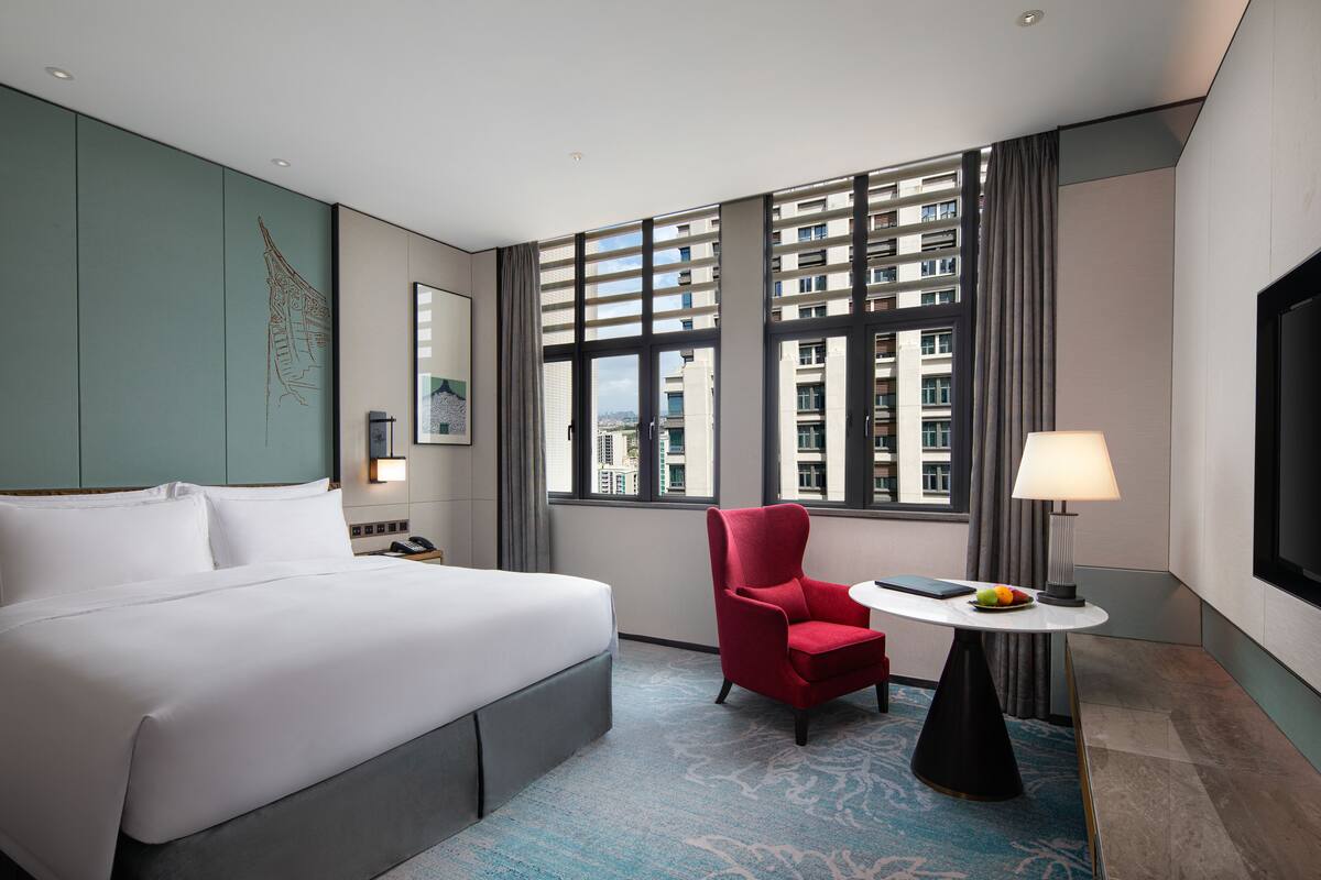 Een hotelkamer met een groot bed, een rode fauteuil, een kleine tafel met een lamp, en een groot raam met uitzicht op de stad.