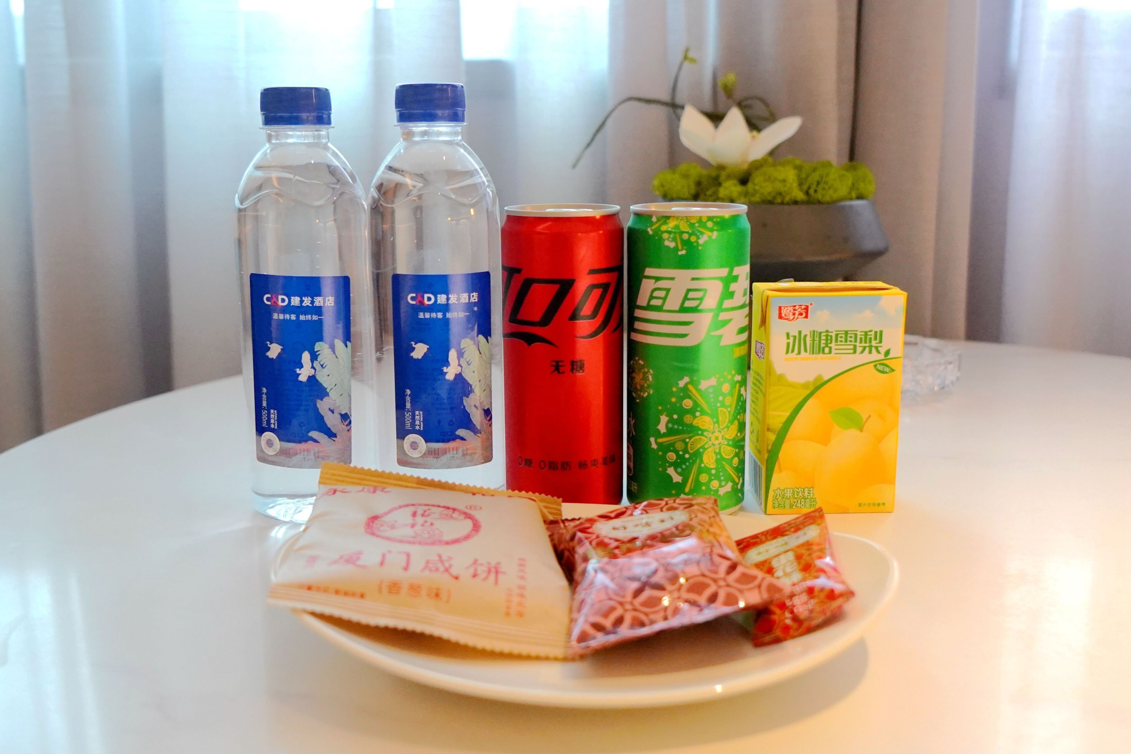 Una mesa con agua embotellada, una lata roja, una lata verde, una caja amarilla y dos paquetes de aperitivos.
