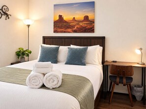 Une chambre d’hôtel comprenant un lit, des oreillers, un bureau, une chaise, une lampe, une décoration murale représentant un paysage désertique et une plante.