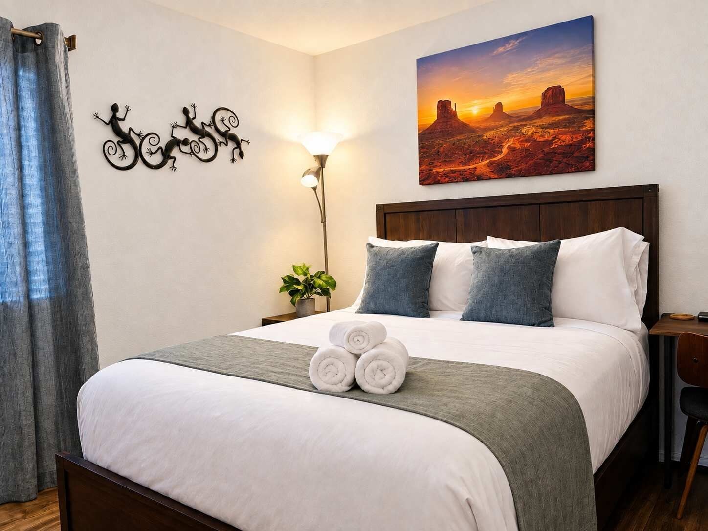 A hotel room with a bed, a nightstand, a lamp, a wall art of a desert sunset, and a decorative wall piece of lizards.