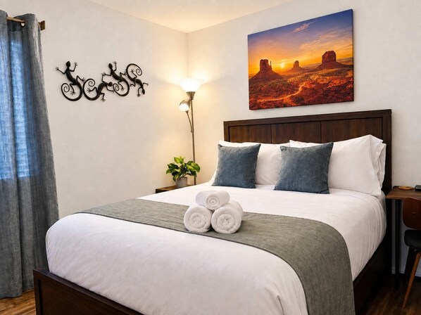 A hotel room with a bed, a nightstand, a lamp, a wall art of a desert sunset, and a decorative wall piece of lizards.