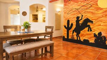 Uma área de refeições com mesa de madeira, bancos e um mural retratando uma cena de deserto com um cavalo e um cavaleiro.