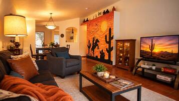 Uma sala de estar com sofá, poltrona, mesa de centro e televisão exibindo uma paisagem desértica.