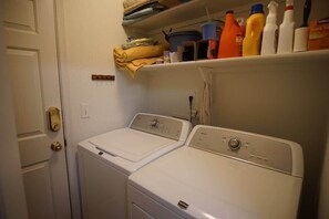 A laundry area with two white washing machines, a shelf with folded towels and cleaning supplies, and a door with a brass handle. - Sonoran Sanctuary - Hot Tub Near Trails & Downtown (Tucson)