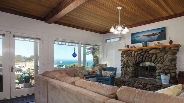 Una sala de estar con chimenea de piedra, un sofá seccional beige grande y vista al océano a través de puertas de cristal.