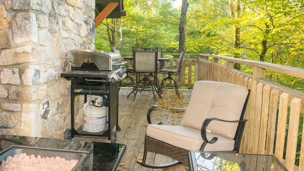 An outdoor patio with a stone wall, a grill, a cushioned chair, and a table. - Misty Mountain Cabin with Grandfather Views (Blowing Rock)