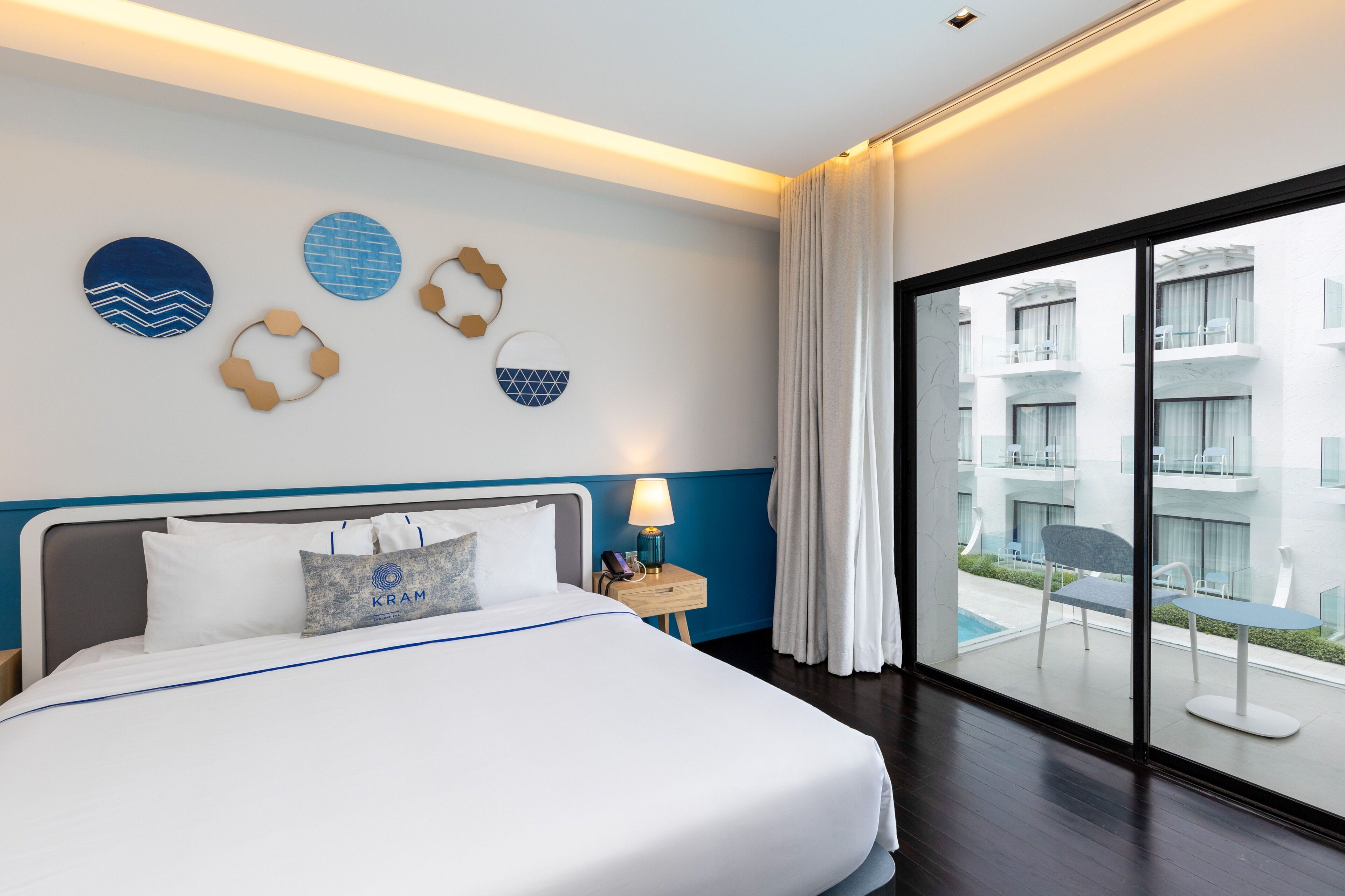 Quarto de hotel com uma cama grande, uma mesinha de cabeceira com abajur e vista para uma piscina e edifícios através de uma janela ampla.