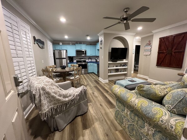 A modern living room with a sofa, a coffee table, and a TV. There is a kitchen area with blue cabinets and a microwave.
