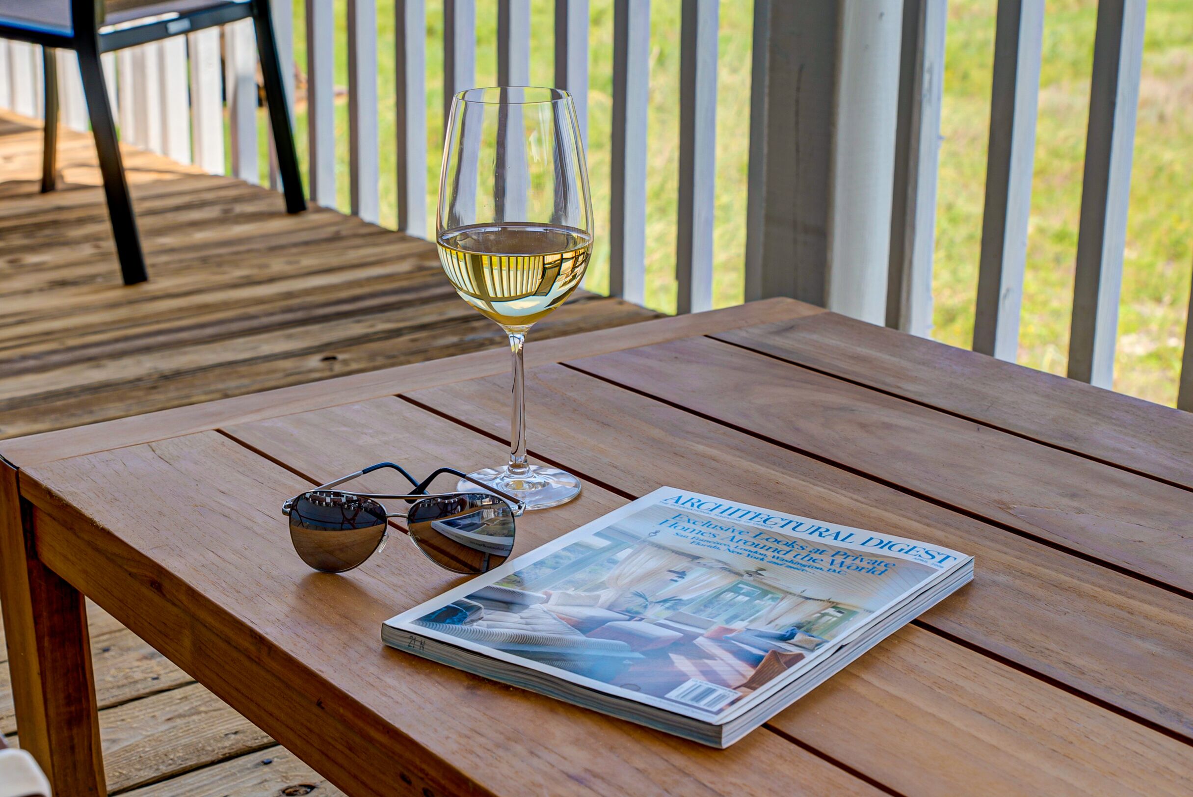 A wooden table with a glass of white wine, a pair of sunglasses, and a magazine titled "ARCHITECTURAL DIGEST".