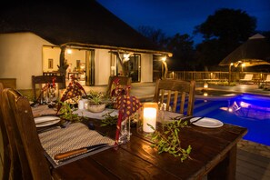 An outdoor dining area with a pool, wooden furniture, and a thatched-roof building. - Vuyani River Lodge (Hoedspruit)
