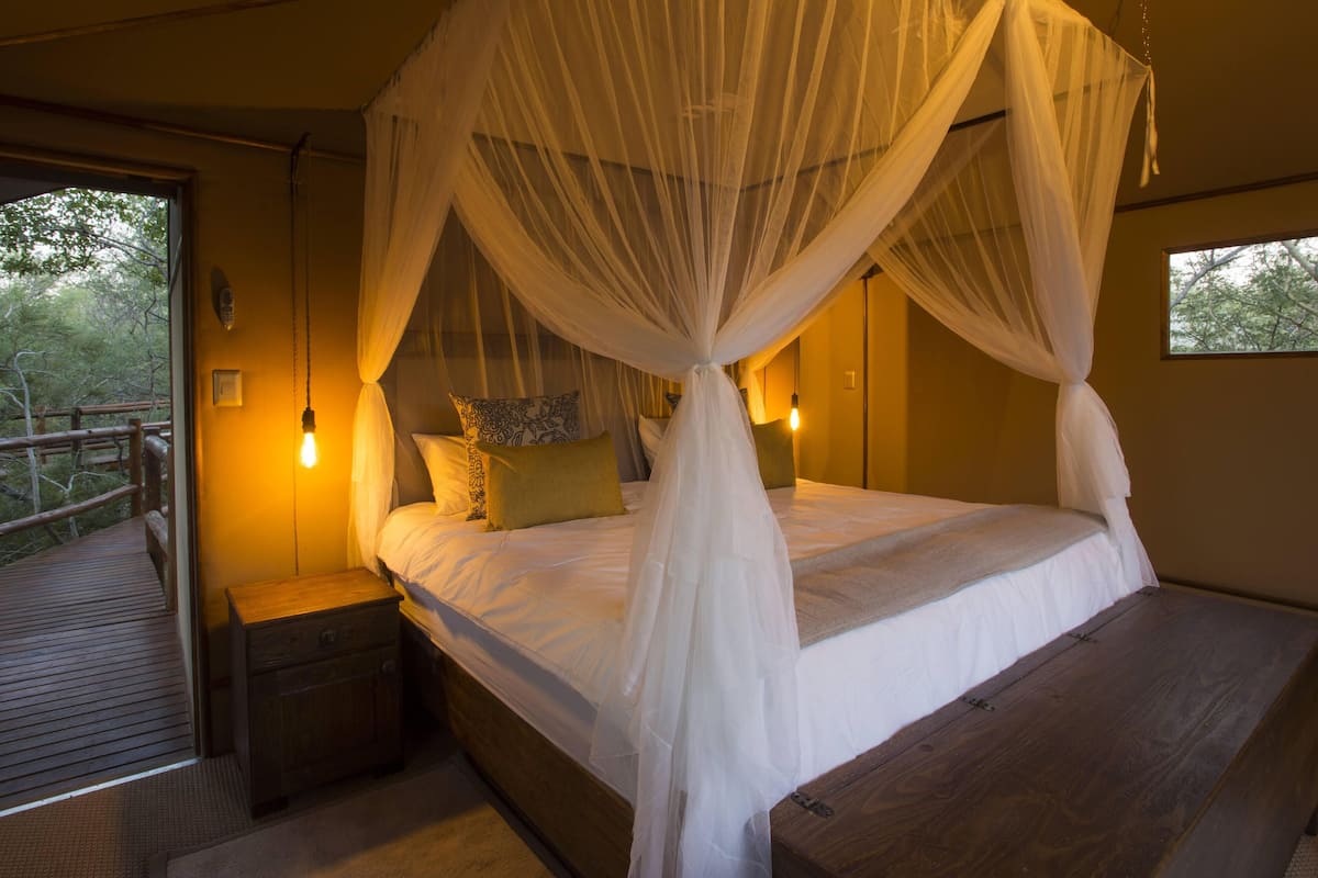 A bedroom with a four-poster bed, a wooden nightstand, and a view of the outdoors.