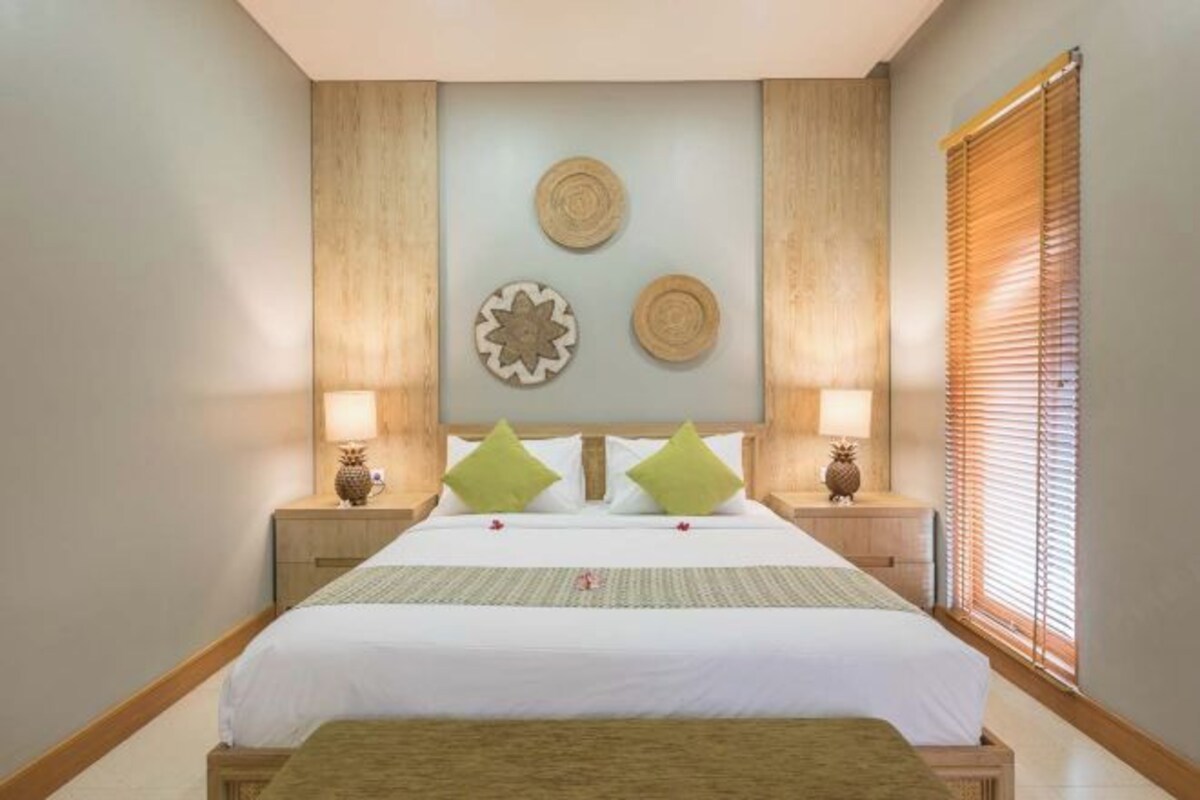 Une chambre d’hôtel avec un grand lit, deux tables de chevet, un banc en bois et une fenêtre avec des stores.