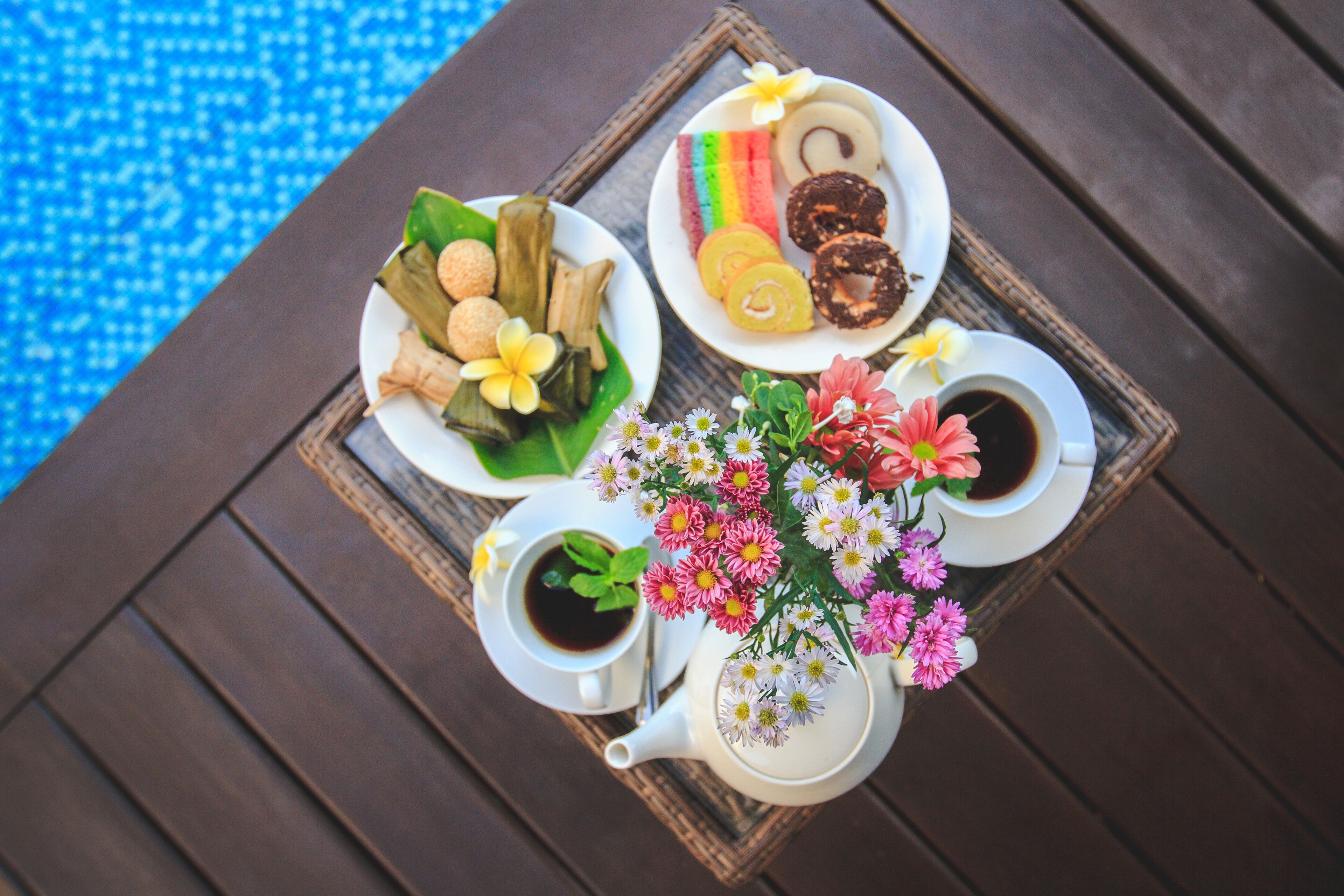 A tray with various food items, a teapot, and a cup of coffee on a wooden table by a pool.