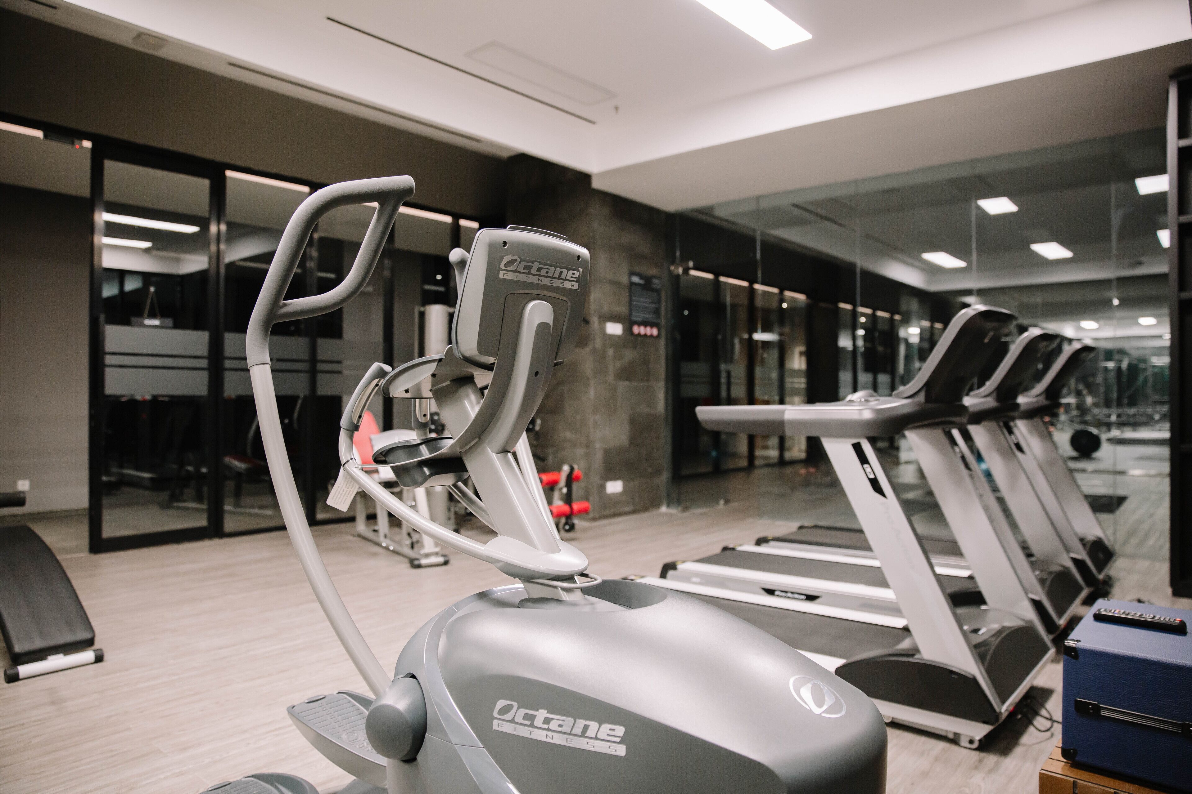 A gym with exercise equipment including treadmills and a stationary bike, with a glass wall and a stone wall in the background.