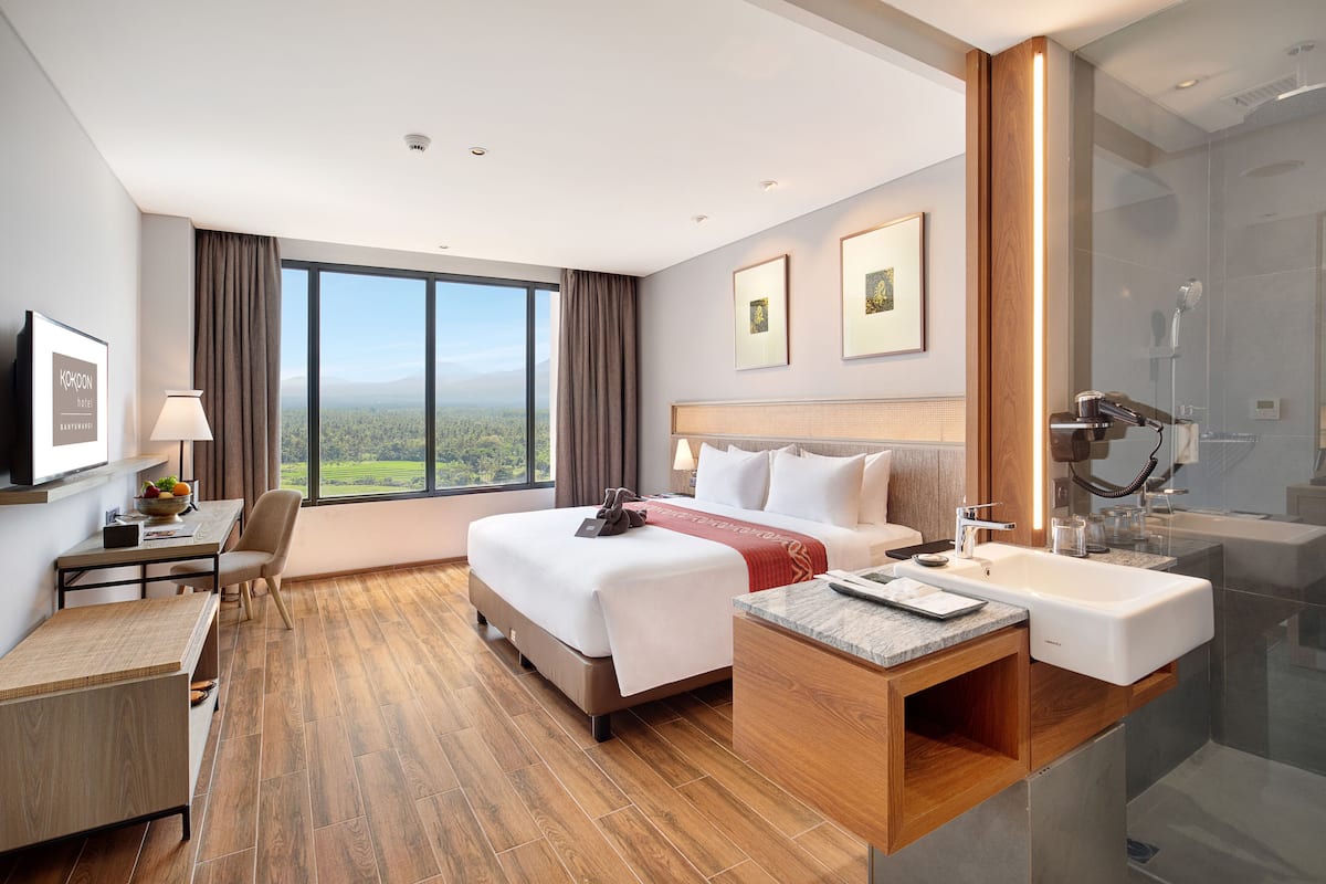 Une chambre d’hôtel dotée d’un grand lit, d’un bureau, d’une chaise, d’une salle de bain avec un lavabo et une douche, et d’une grande fenêtre offrant une vue sur le paysage.