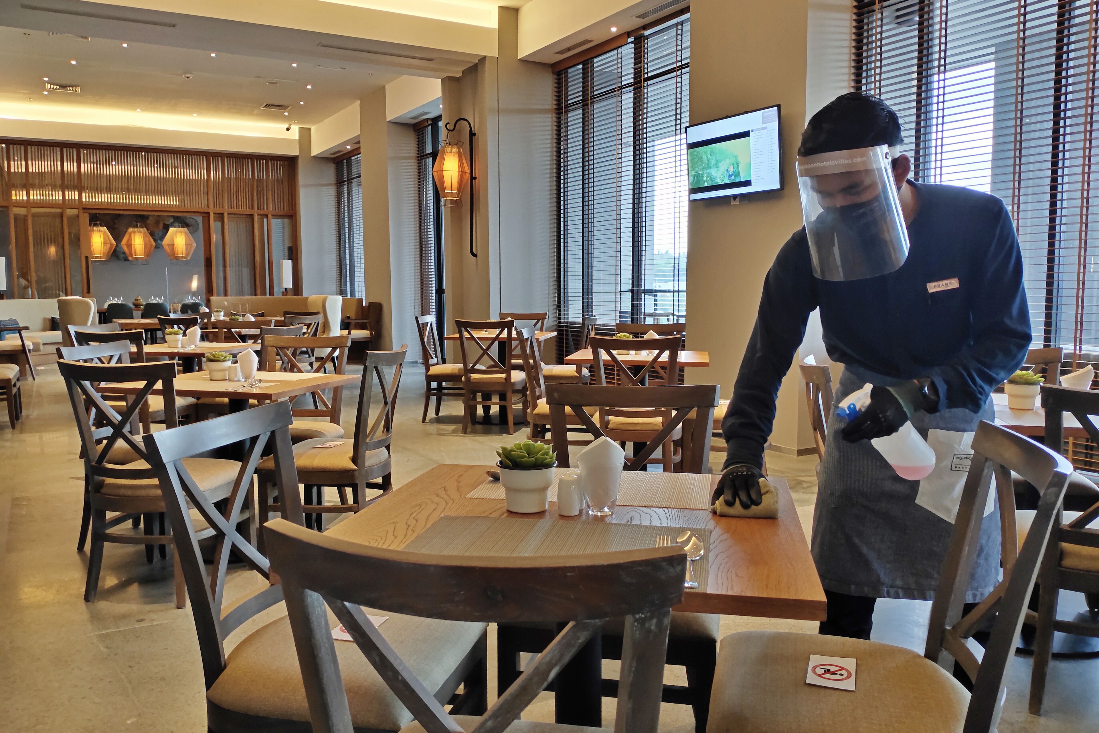 A restaurant with wooden tables and chairs, a TV screen on the wall, and a person cleaning a table.
