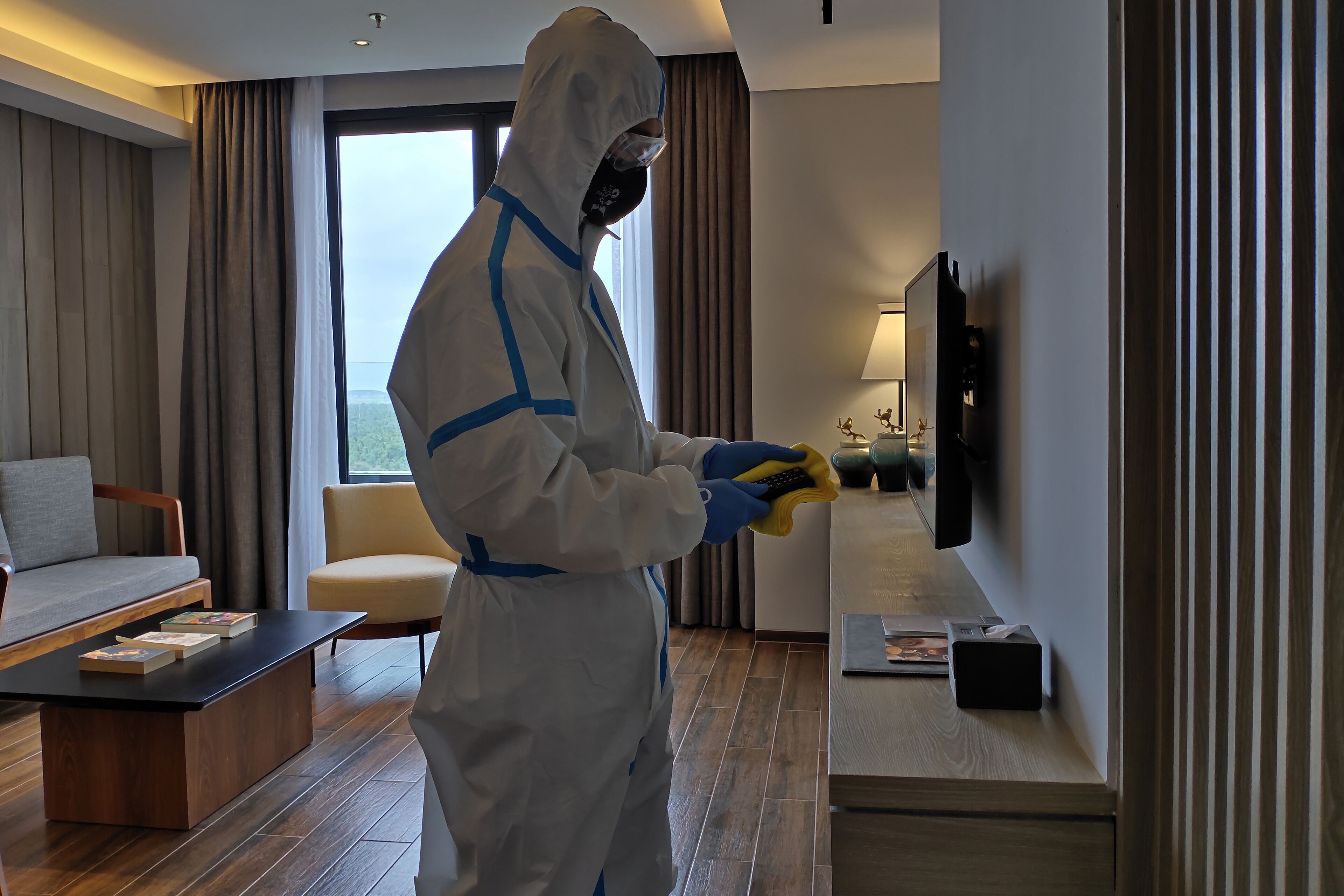 Eine Person in Schutzkleidung reinigt einen Fernseher in einem modernen Hotelzimmer, das eine Sitzecke, einen Sessel und einen Blick ins Freie bietet.