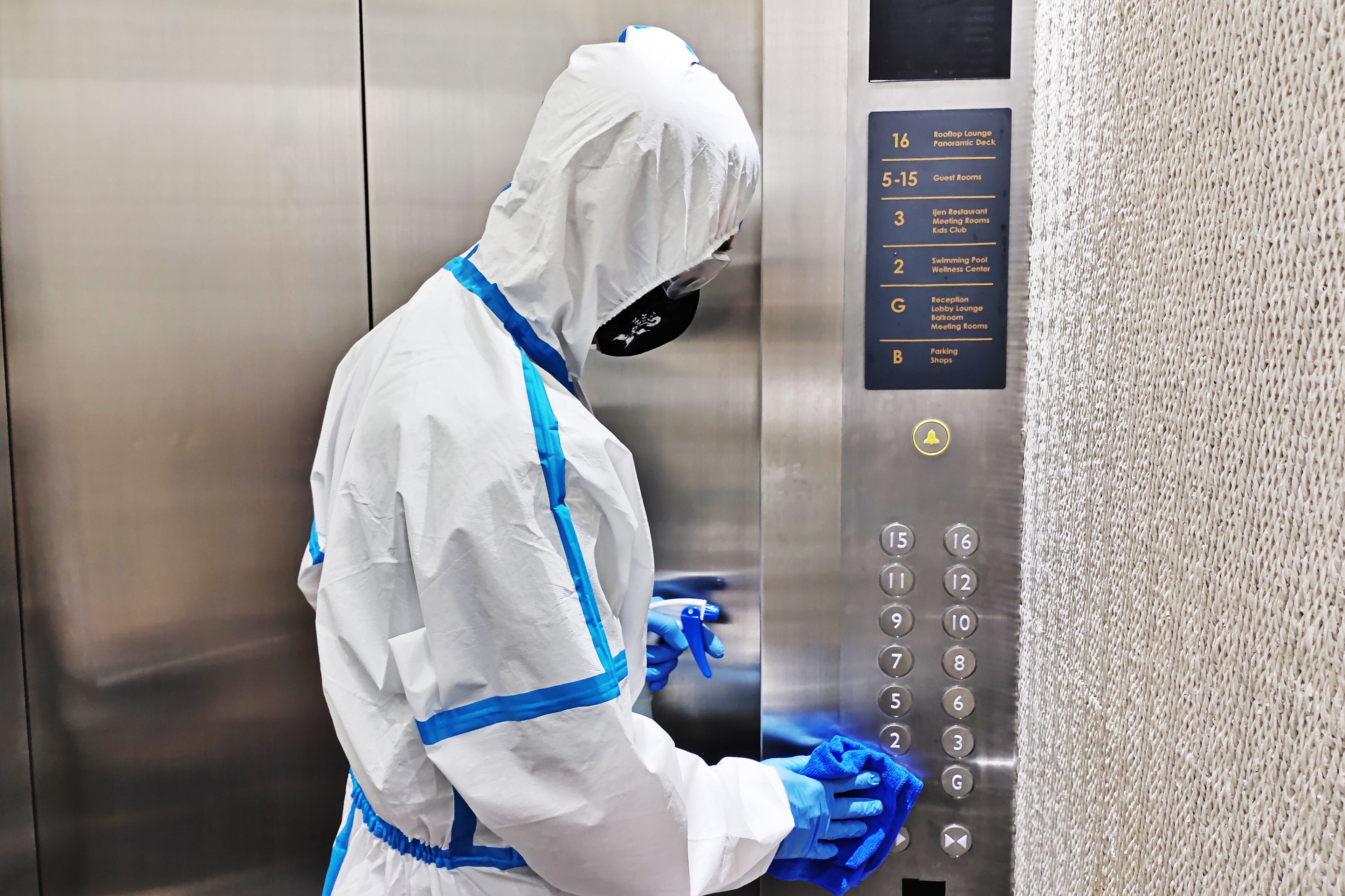 A person in a white protective suit and blue gloves operating an elevator control panel with numbered buttons and a list of room types.