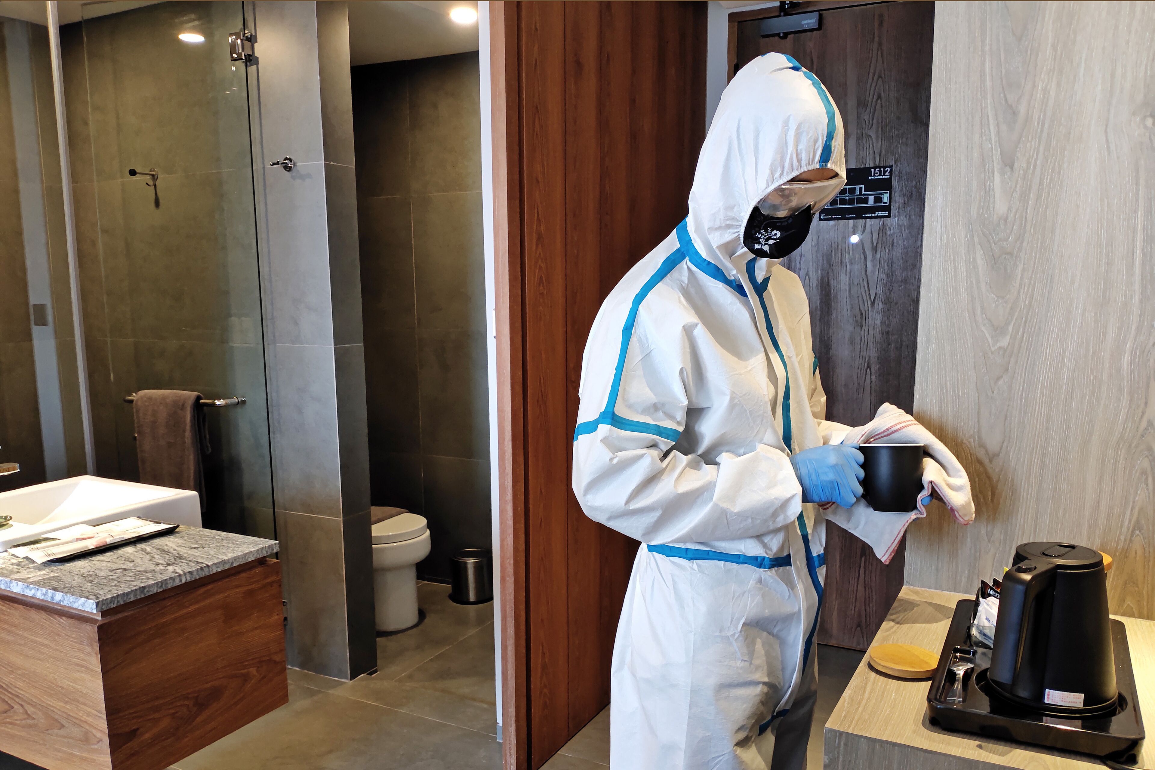 A person in a white protective suit with blue stripes, holding a black cup and a cloth, standing in a modern bathroom with a wooden vanity, a glass shower enclosure, and a wooden door.