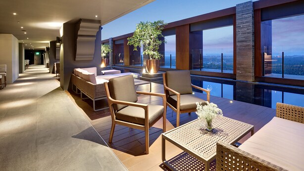 A modern outdoor seating area with wooden furniture and large windows overlooking a cityscape.