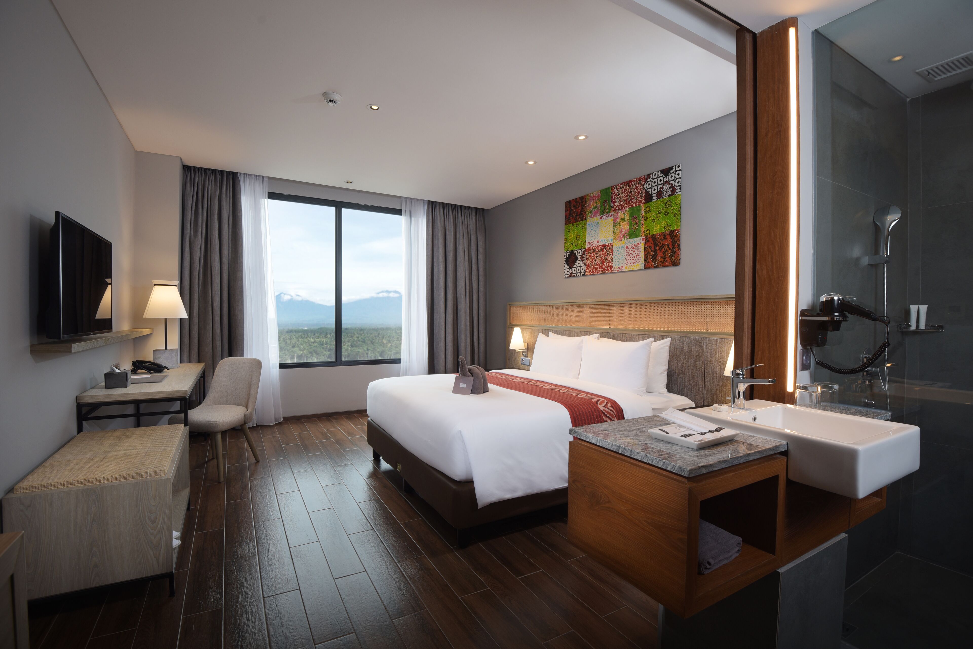 A modern hotel room with a large bed, a desk, a chair, a bathroom with a sink and mirror, and a view of the mountains.