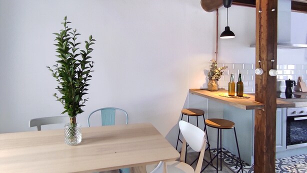 Un coin repas avec une table en bois, des chaises blanches, un luminaire suspendu et une cuisine en arrière-plan.