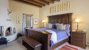 A bedroom with a large bed, wooden headboard, two bedside tables with lamps, a bench, a fireplace, and a painting on the wall. - Stylish and Serene, Community Pool (Santa Fe)