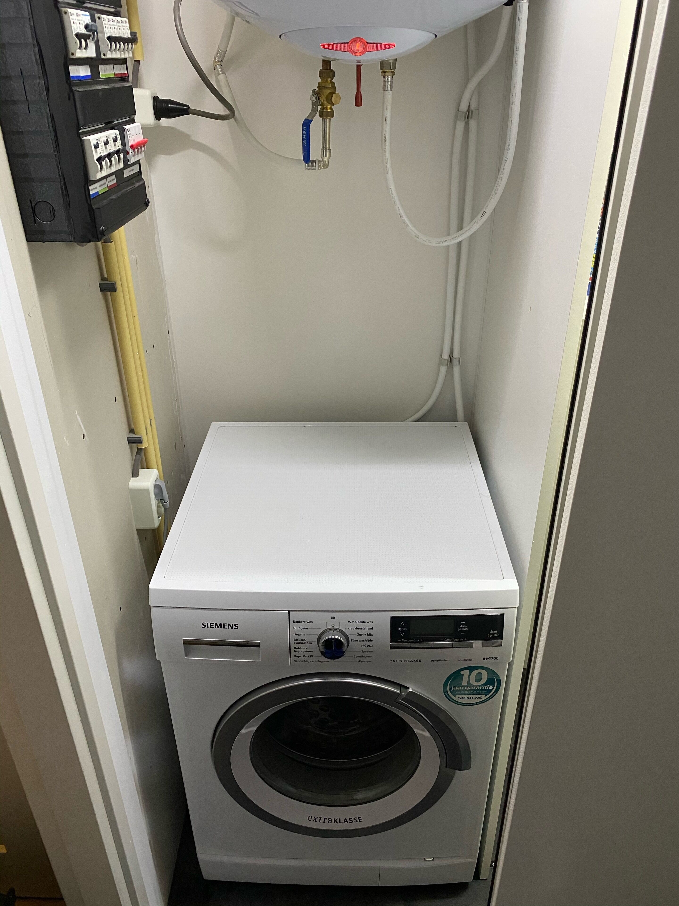 A compact, white Siemens washing machine with a front-loading door, located in a small laundry room with a white ceiling and visible plumbing.