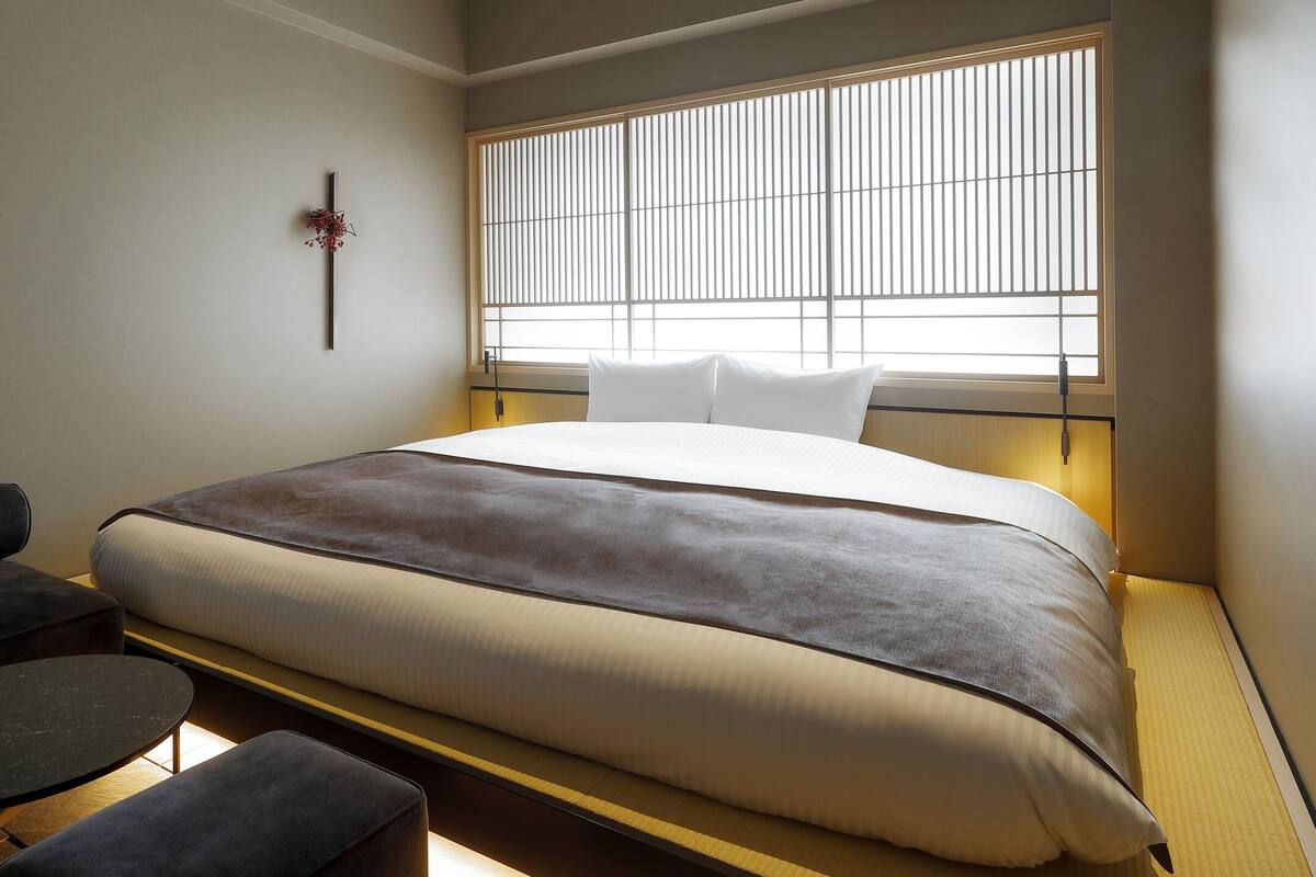 Una habitación de hotel con una cama grande, una mesita de noche, una silla y una ventana con persianas verticales.