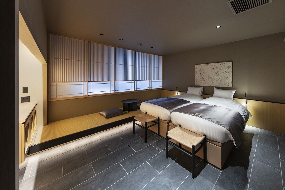 Un dormitorio moderno con cama, mesitas de noche, una ventana con persianas, una obra de arte colgada en la pared y un banco empotrado con espacio de almacenamiento.